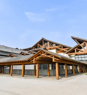 Exterior view of a large building with multiple overlapping pitched roofs and exposed timber trusses, blending traditional architectural elements with modern materials. The structure is surrounded by paved open space and landscaping.