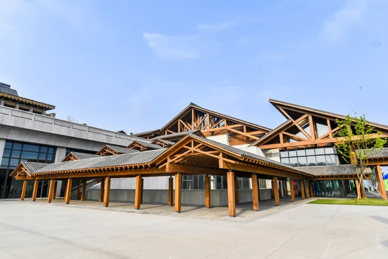 Exterior view of a large building with multiple overlapping pitched roofs and exposed timber trusses, blending traditional architectural elements with modern materials. The structure is surrounded by paved open space and landscaping.
