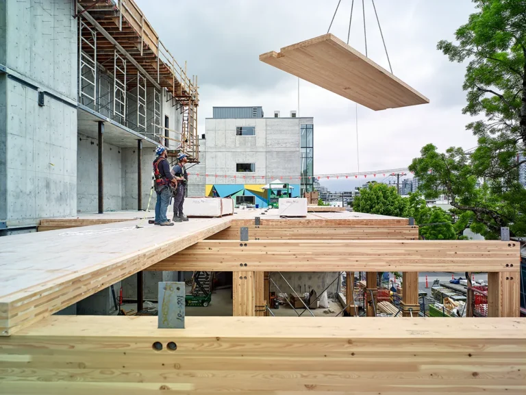 Construction workers in safety gear stand on a mass timber structure, guiding a large wooden panel being lifted by a crane. The site features exposed timber beams, scaffolding, and concrete walls, with urban buildings and trees visible in the background.