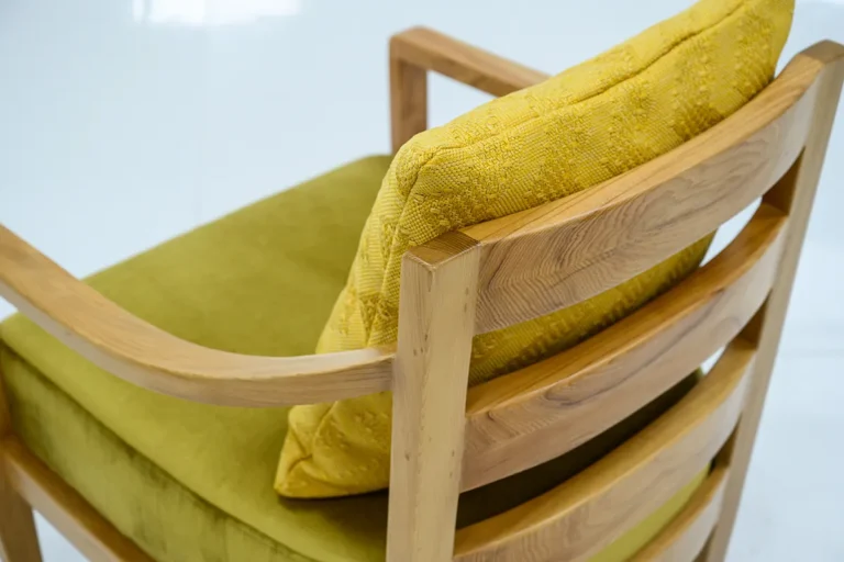Close-up view of a wooden chair with curved armrests and back slats, featuring a green seat cushion and a textured yellow pillow.