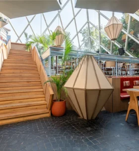 nterior of a bright, stylish restaurant or café featuring a central wooden staircase, geometric pendant lights, potted plants, and a mix of seating including a red banquette and wooden chairs, set beneath a glass atrium ceiling.