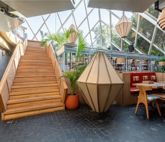 nterior of a bright, stylish restaurant or café featuring a central wooden staircase, geometric pendant lights, potted plants, and a mix of seating including a red banquette and wooden chairs, set beneath a glass atrium ceiling.
