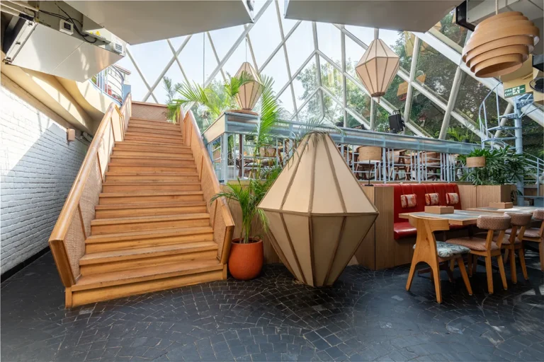 nterior of a bright, stylish restaurant or café featuring a central wooden staircase, geometric pendant lights, potted plants, and a mix of seating including a red banquette and wooden chairs, set beneath a glass atrium ceiling.