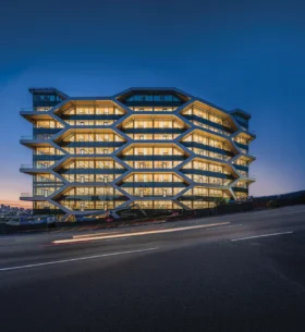Exterior of a multi-storey office building at dusk, featuring a distinctive geometric façade with large windows illuminated from within.