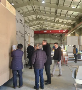 Visitors tour a prefabricated wood building manufacturing facility, examining a modular wall assembly while standing on the production floor surrounded by construction materials and equipment.