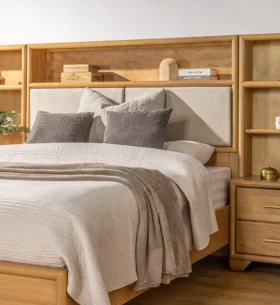 Cozy bedroom featuring a light wood bed frame with an upholstered headboard and integrated shelving on either side. The shelves are decorated with books, vases, plants, and small decor items. The bed is dressed in neutral bedding with soft gray and beige pillows and a throw blanket, set against a light-coloured wall and wood flooring.