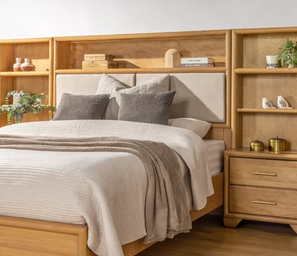 Cozy bedroom featuring a light wood bed frame with an upholstered headboard and integrated shelving on either side. The shelves are decorated with books, vases, plants, and small decor items. The bed is dressed in neutral bedding with soft gray and beige pillows and a throw blanket, set against a light-coloured wall and wood flooring.