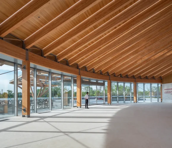 Spacious interior of a mass timber building with exposed wooden beams, floor-to-ceiling glass walls, and abundant natural light casting shadows on the floor. A person stands near the windows, and trees are visible outside.