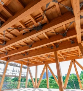 Interior view of a mass timber structure under construction, featuring exposed wooden beams, columns, and steel connectors. Scaffolding is visible along the windows, and the space is filled with natural light from large openings that reveal green trees and a clear sky outside.
