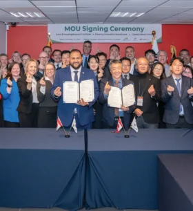 Participants gather behind two representatives holding signed agreements during a memorandum of understanding signing ceremony, with Canadian and Korean flags displayed on a table beneath an event banner.