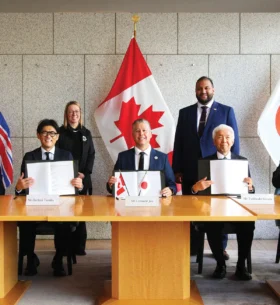Representatives from Canadian and Japanese organizations seated at a conference table holding signed documents, with Canadian and Japanese flags displayed behind them during a memorandum of understanding signing ceremony.