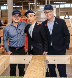 Three visitors wearing branded caps stand beside a wood-framed structural wall assembly inside a manufacturing facility, with engineered wood components and production equipment visible in the background.