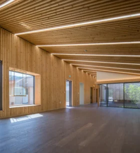 Spacious, modern interior with angled mass timber ceiling and walls clad in vertical wooden slats. Large windows and glass doors let in abundant natural light, offering views of greenery and an outdoor area. The room features sleek, integrated lighting and a minimalist aesthetic.