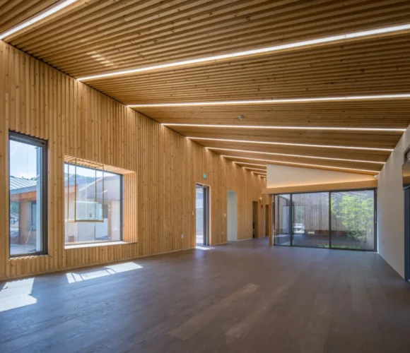 Spacious, modern interior with angled mass timber ceiling and walls clad in vertical wooden slats. Large windows and glass doors let in abundant natural light, offering views of greenery and an outdoor area. The room features sleek, integrated lighting and a minimalist aesthetic.