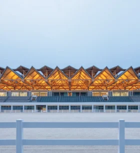 An equestrian centre featuring an illuminated mass timber roof structure with exposed wood beams and repeating triangular forms above spectator seating.
