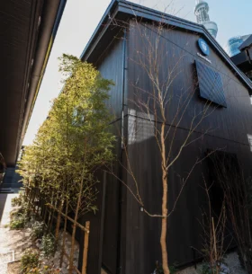 Exterior of a black wood-clad building at the T-home KEI Hotel in Tokyo, framed by landscaped trees and shrubs.