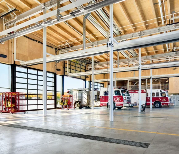 Interior view of a fire station with large glass bay doors and extensive use of exposed structural wood for the roof and walls.