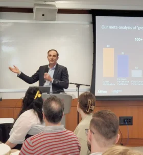 A speaker presents at a conference while standing at a podium and holding a microphone. Attendees are seated in the foreground, facing a projection screen displaying a slide with a bar chart about green building certifications and performance standards.