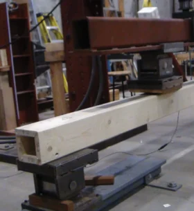 A hollow wood beam mounted on a structural testing machine in a laboratory. The light-coloured beam rests on steel supports while a hydraulic loading frame applies pressure from above. Ladders, equipment and workbenches are visible in the background.