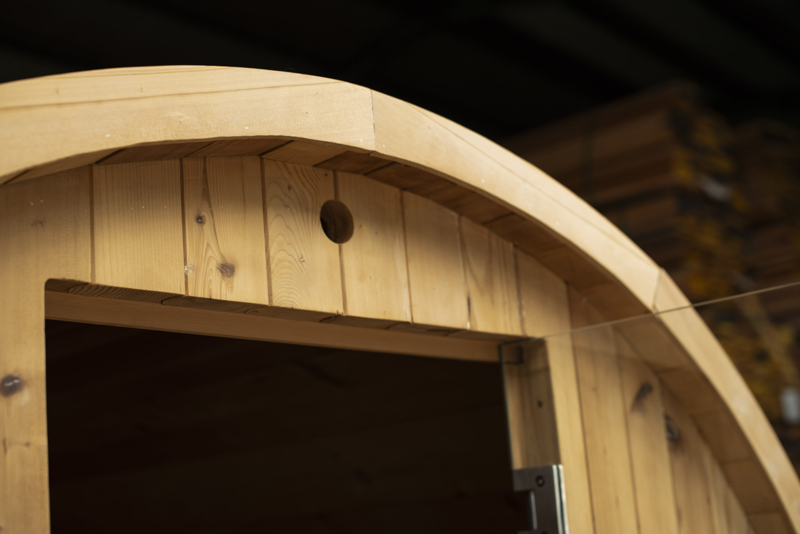 A close-up shot shows the curved top and doorway of a thermally modified hemlock sauna. The light-colored wood has vertical panels and a smooth, rounded arch forming the roof and the top of the doorframe. A small, round ventilation hole is visible near the peak of the arch. The interior of the sauna is dark and out of focus. In the blurred background, stacks of lumber are visible.