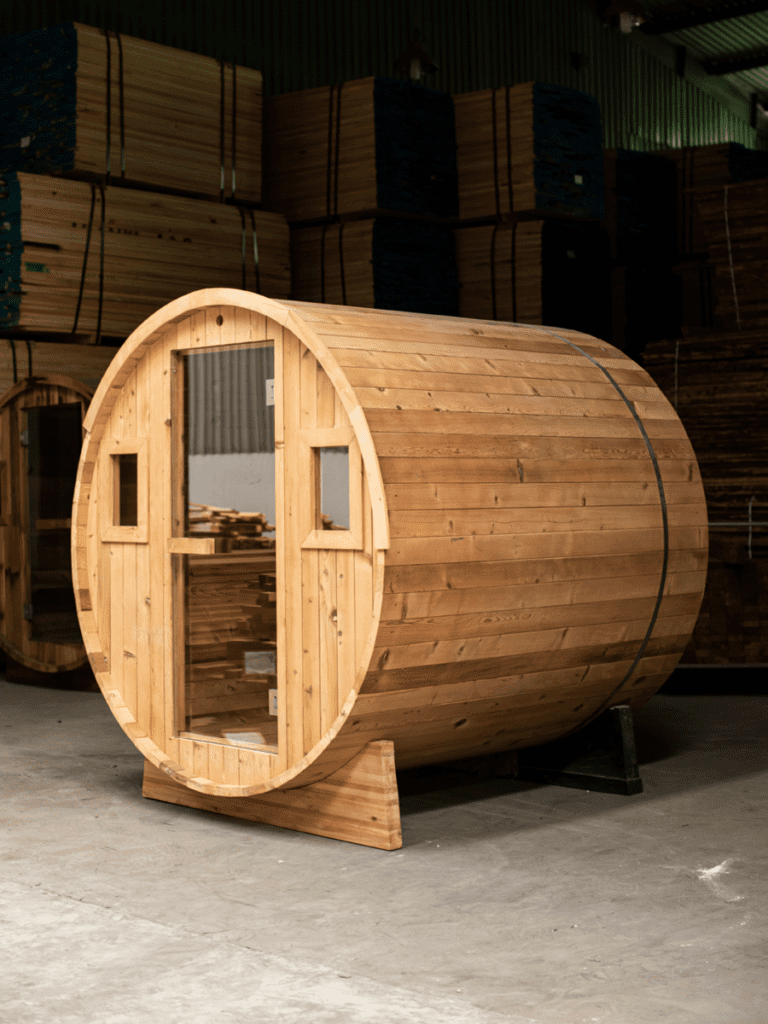 A thermally modified hemlock barrel sauna sits indoors, showcasing its cylindrical shape and light-colored wood construction with horizontal slats. The sauna features a glass-paneled door in the center of one circular end and two small, square windows flanking it. Metal bands encircle the barrel for stability. It rests on wooden supports. The background shows a warehouse setting with stacks of lumber.