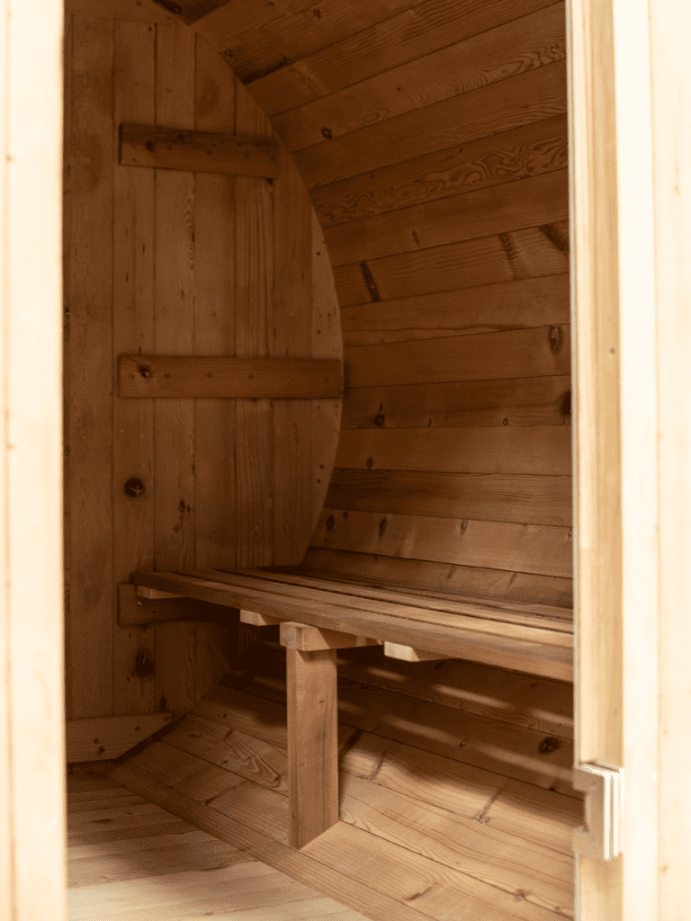 The interior of a thermally modified hemlock barrel sauna is shown from the doorway. Light-colored wooden planks line the curved walls and ceiling, as well as the floor. A single wooden bench with slats runs along the back curved wall, supported by sturdy wooden legs. A small, dark circular vent is visible on the side wall. The doorframe is in the foreground, slightly out of focus.
