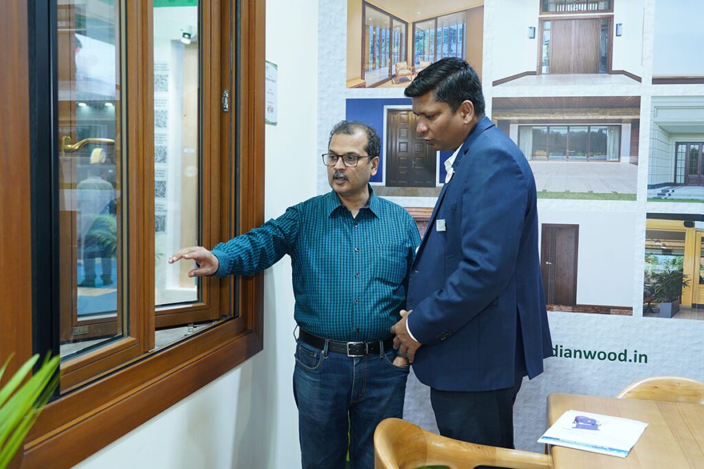 Two men are standing next to a large wooden-framed window, with one man pointing towards the window as if explaining something, at the Canadian Wood booth.