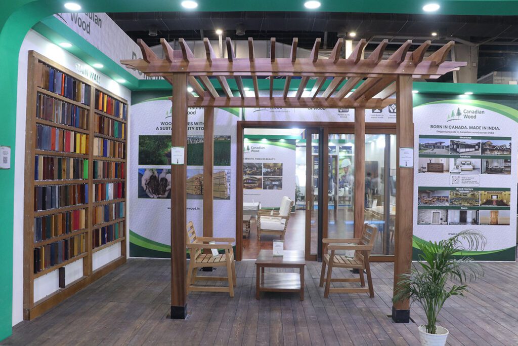 A wide shot of the Canadian Wood booth at IndiaWood 2025, featuring a wooden pergola with two chairs and a table, a wall display of colorful wood samples, and informational posters on the right.