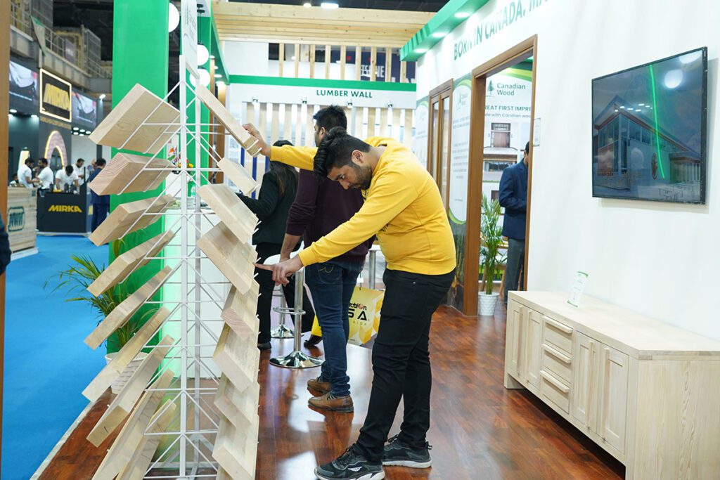 A man in a yellow sweatshirt inspects a display of various wood lumber samples arranged horizontally on white metal racks at the Canadian Wood booth.