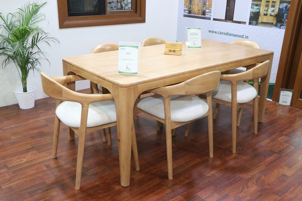 A close-up of a light-colored wooden dining table with six matching chairs with white upholstered seats, displayed in a showroom setting.