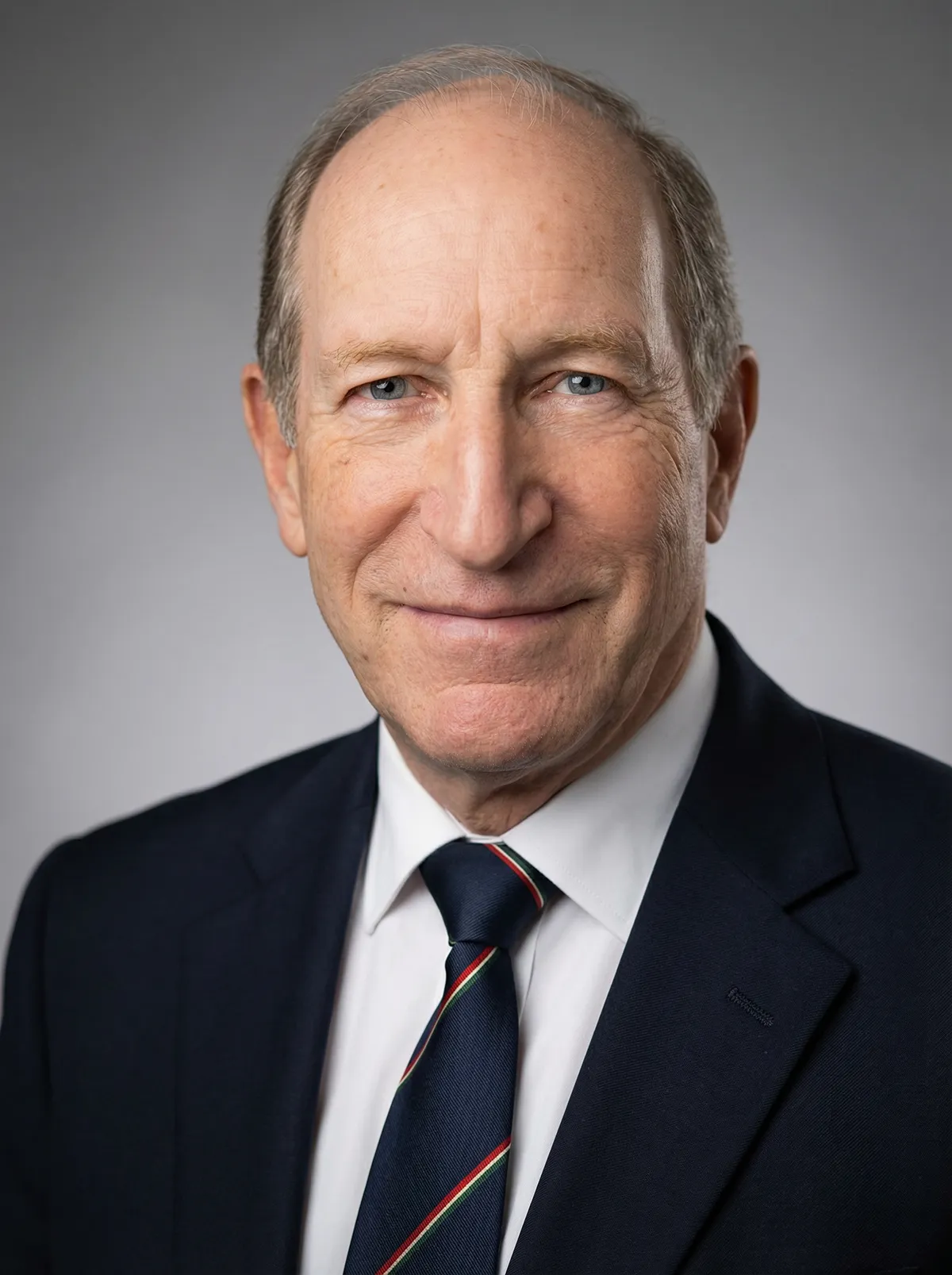 A headshot of an older man with light skin, blue eyes, and thin, receding light-brown hair. He is wearing a dark navy blue suit jacket, a crisp white dress shirt, and a navy blue necktie featuring thin, diagonal red and green stripes. He has a slight, pleasant smile and is looking directly at the camera against a soft, neutral grey gradient background.