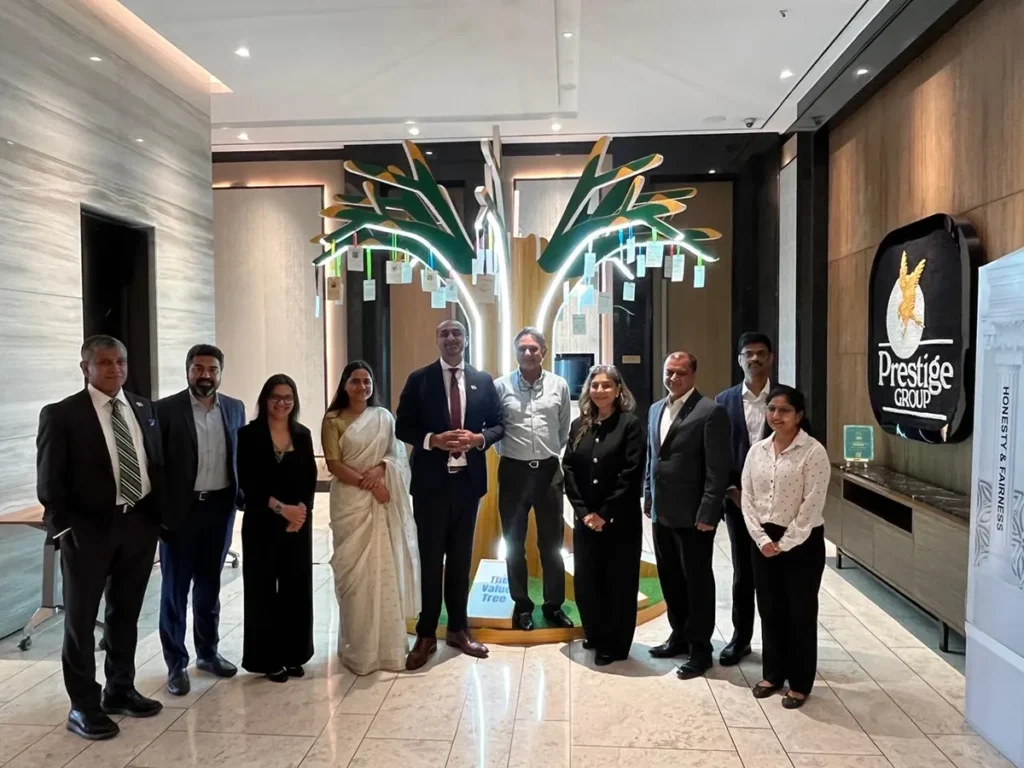 Minister Ravi Kahlon and a delegation posing indoors with representatives of the Prestige Group in India. They are standing in a modern, bright lobby in front of a decorative, glowing tree art installation. To the right, the Prestige Group logo is visible on a dark wall panel.
