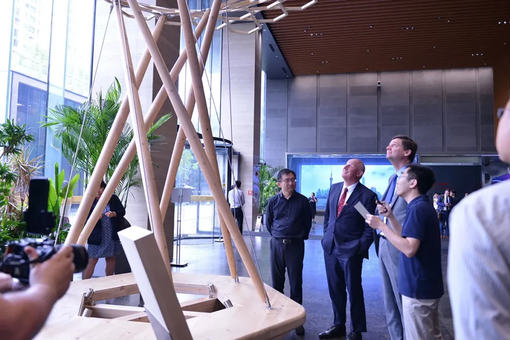 Premier David Eby and delegation members view a large timber structural installation inside the ECADI building, looking up as representatives explain the wood design and construction features.