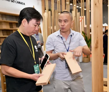 An FII Vietnam staff member discusses wood product samples with an attendee at a Canadian Wood exhibition booth, comparing boards and explaining material characteristics.