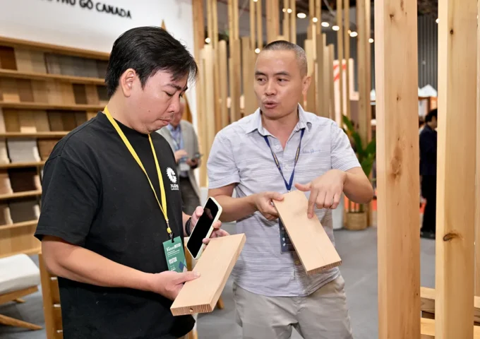An FII Vietnam staff member discusses wood product samples with an attendee at a Canadian Wood exhibition booth, comparing boards and explaining material characteristics.