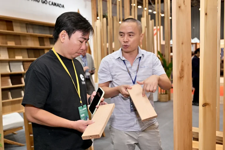 An FII Vietnam staff member discusses wood product samples with an attendee at a Canadian Wood exhibition booth, comparing boards and explaining material characteristics.