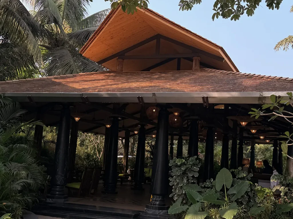 Open-air pavilion with a prominent wood shingle roof, exposed timber ceiling and wood architectural details set within a tropical landscape.